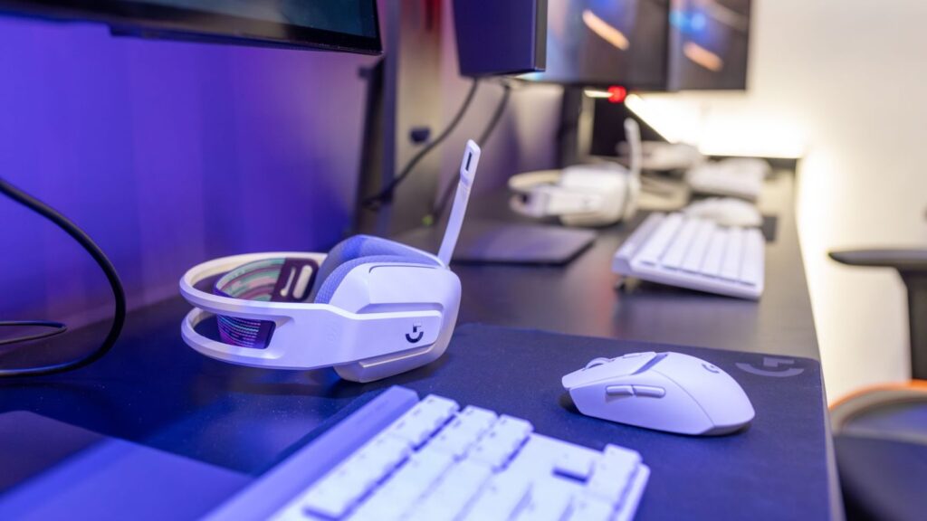 Juodos žaidimų ausinės su mikrofonu ant stalo prie klaviatūros, šviesa iš monitoriaus