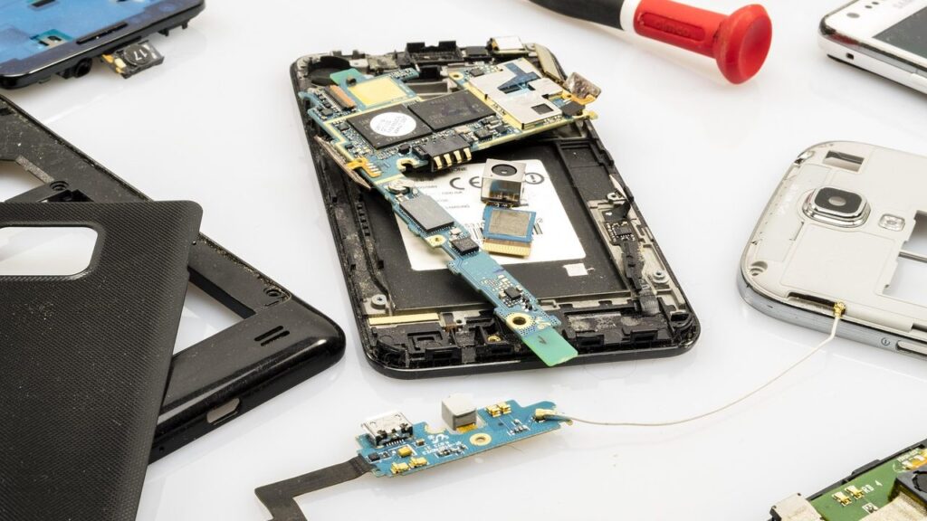 Elektronikos remonto dirbtuvių darbo stalas su išardytu telefonu ir laukimo laikrodis fone